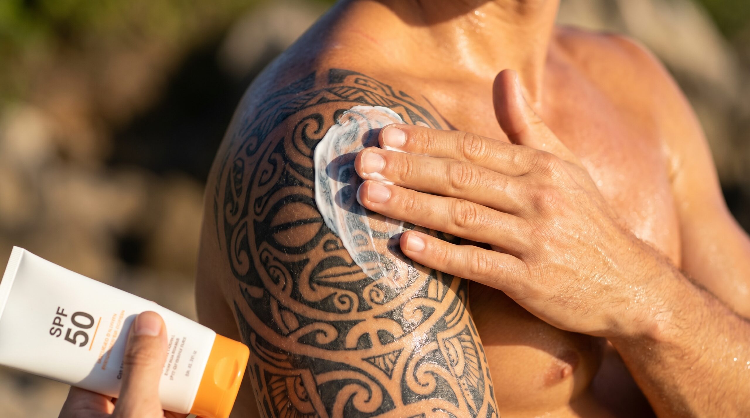 Fotografia ravvicinata lifestyle di una mano che applica uniformemente una crema solare ad alta protezione (SPF 50) su un grande tatuaggio tribale scuro sulla spalla all’aperto, con la nitida luce del mattino che illumina la pelle e mostra la corretta preservazione a lungo termine e l’idratazione del body art nero intenso.