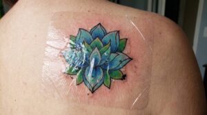 Fotografia ravvicinata di un tatuaggio fresco e vibrante su una spalla, coperto da una benda medica trasparente professionale "effetto seconda pelle". Si notano minuscole gocce di plasma e inchiostro in eccesso intrappolate sotto la pellicola. La pelle circostante è leggermente rosa e infiammata.