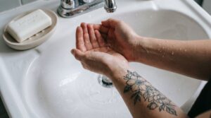 Una ripresa in prima persona sopra un lavandino del bagno. Delle mani tengono a coppa dell'acqua tiepida per sciacquare delicatamente un tatuaggio appena fatto. Una saponetta neutra e antibatterica è visibile di lato. L'immagine dovrebbe trasmettere un senso di delicatezza, pulizia e cura.