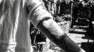 Fotografia ad alto contrasto del braccio di una persona con un tatuaggio appena fatto, parzialmente ombreggiato da una leggera camicia di lino. Metà del tatuaggio è nascosta dal tessuto, mentre l'altra metà è colpita da un raggio di sole forte e abbagliante, a rappresentare visivamente la fragilità dell'inchiostro. Sullo sfondo si vede una luminosa terrazza estiva.