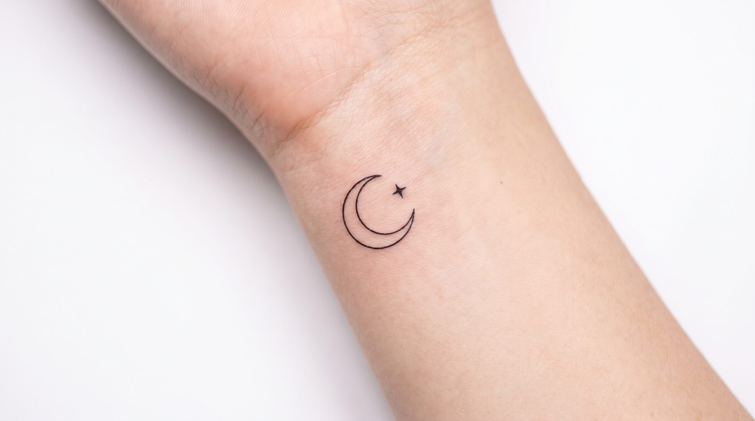 Foto macro di un micro tatuaggio impeccabile all’interno del polso, con una minuscola luna crescente fine-line e una piccola stella, inferiore a due centimetri, dai bordi nitidi, inchiostro definito e sfondo bianco pulito.