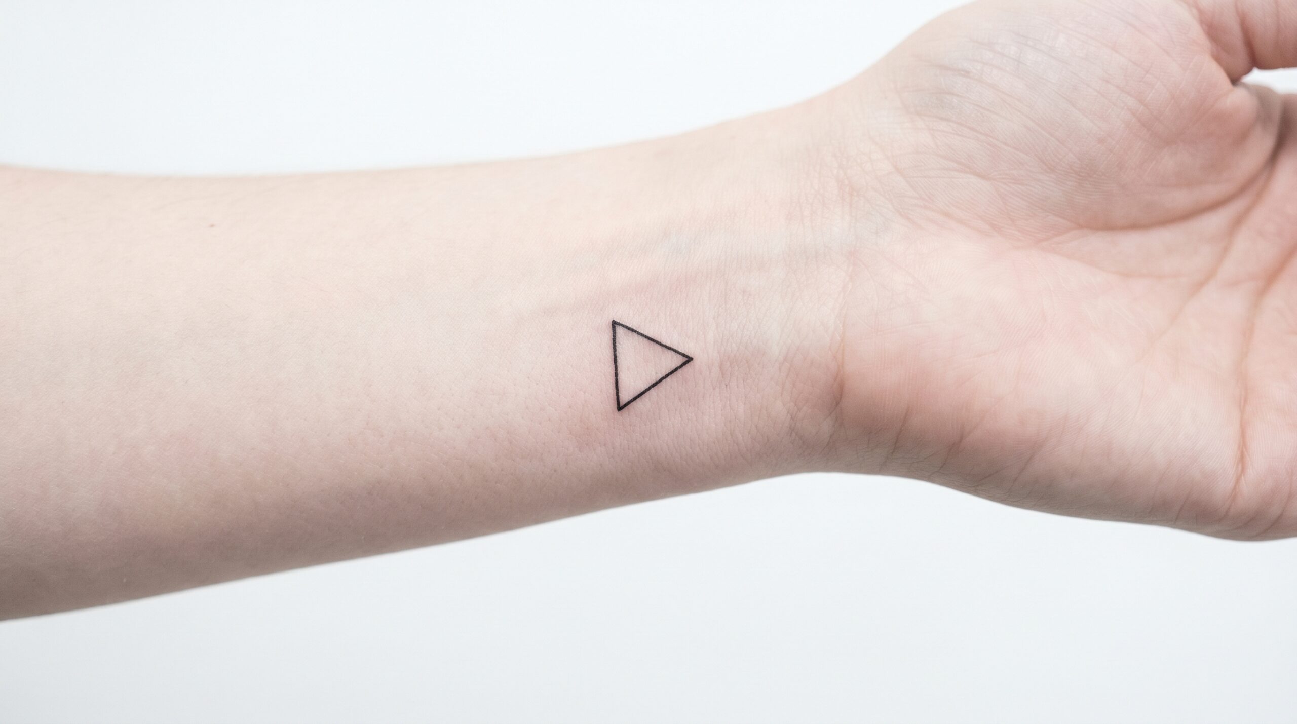 Tatuaggio minimalista di un piccolo triangolo aperto e nitido sul bordo esterno del polso, con linee pulite, allineamento anatomico bilanciato, estetica delicata e moderna, isolato su sfondo bianco.