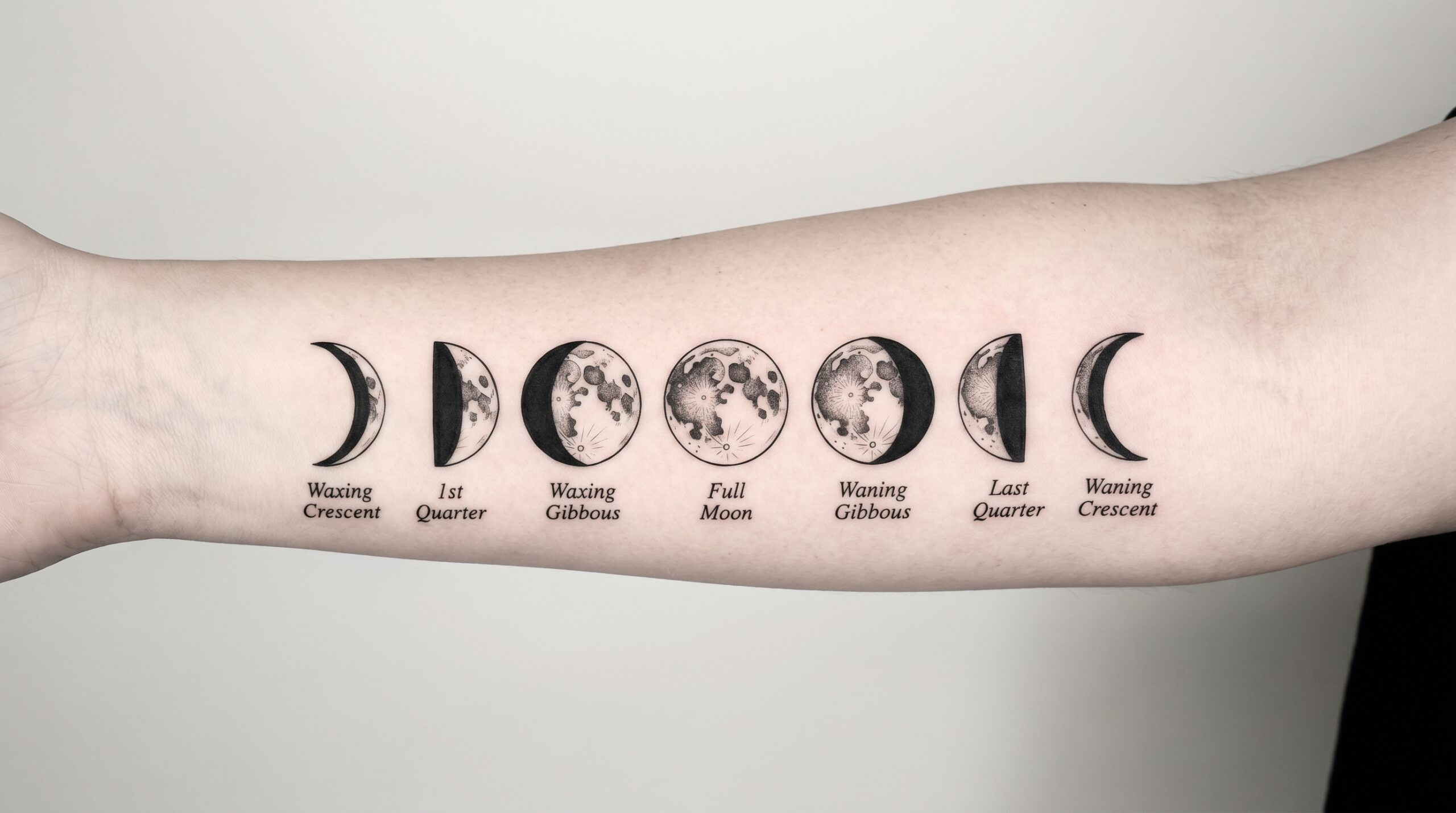 Layout di tatuaggio minimalista che mostra la progressione orizzontale delle 7 principali fasi lunari, dalla sottile luna crescente fino alla luna piena e poi alla luna calante. Forme nere pulite e compatte, arricchite da linework estremamente fine e dettagliato.