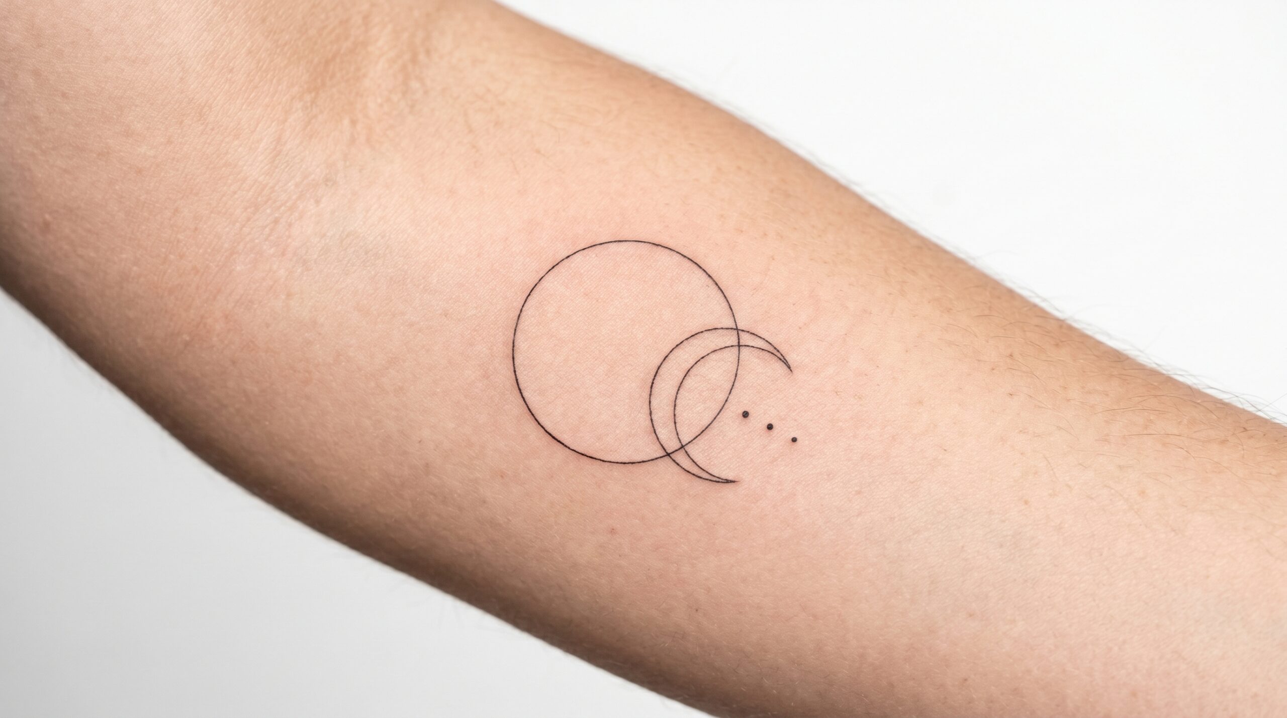 Foto macro in studio di un elegante tatuaggio geometrico fine-line sull’interno dell’avambraccio, con un cerchio sottile che interseca una delicata luna crescente e tre micro punti, illuminazione brillante e dettagli ultra nitidi.