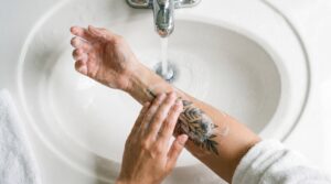 Una persona sta lavando delicatamente un nuovo tatuaggio sull'avambraccio nel lavandino del bagno. Utilizza la punta delle dita con movimenti circolari e un sapone delicato, bianco e schiumoso. L'acqua tiepida scorre dolcemente.