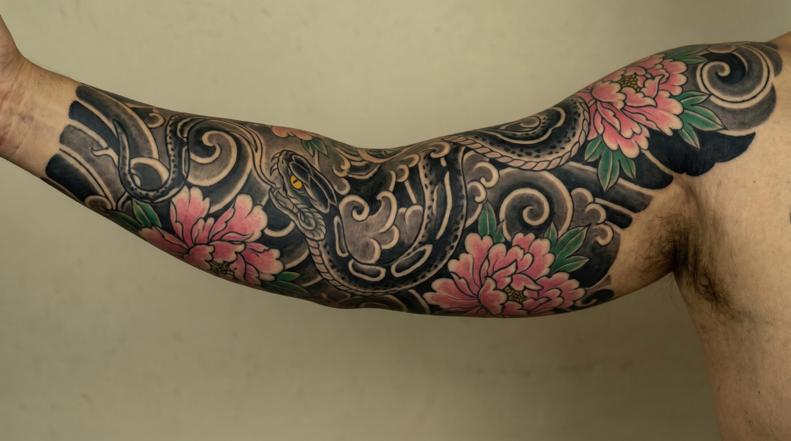 "Design di tatuaggio giapponese Irezumi per manica completa raffigurante un potente serpente Hebi in stile black and grey con intensi occhi gialli, avvolto tra vivaci fiori di peonia rosa e onde di vento stilizzate scure. Linee marcate e pulite, ombreggiature nere solide e autentico stile artistico del periodo Edo ad alto contrasto."